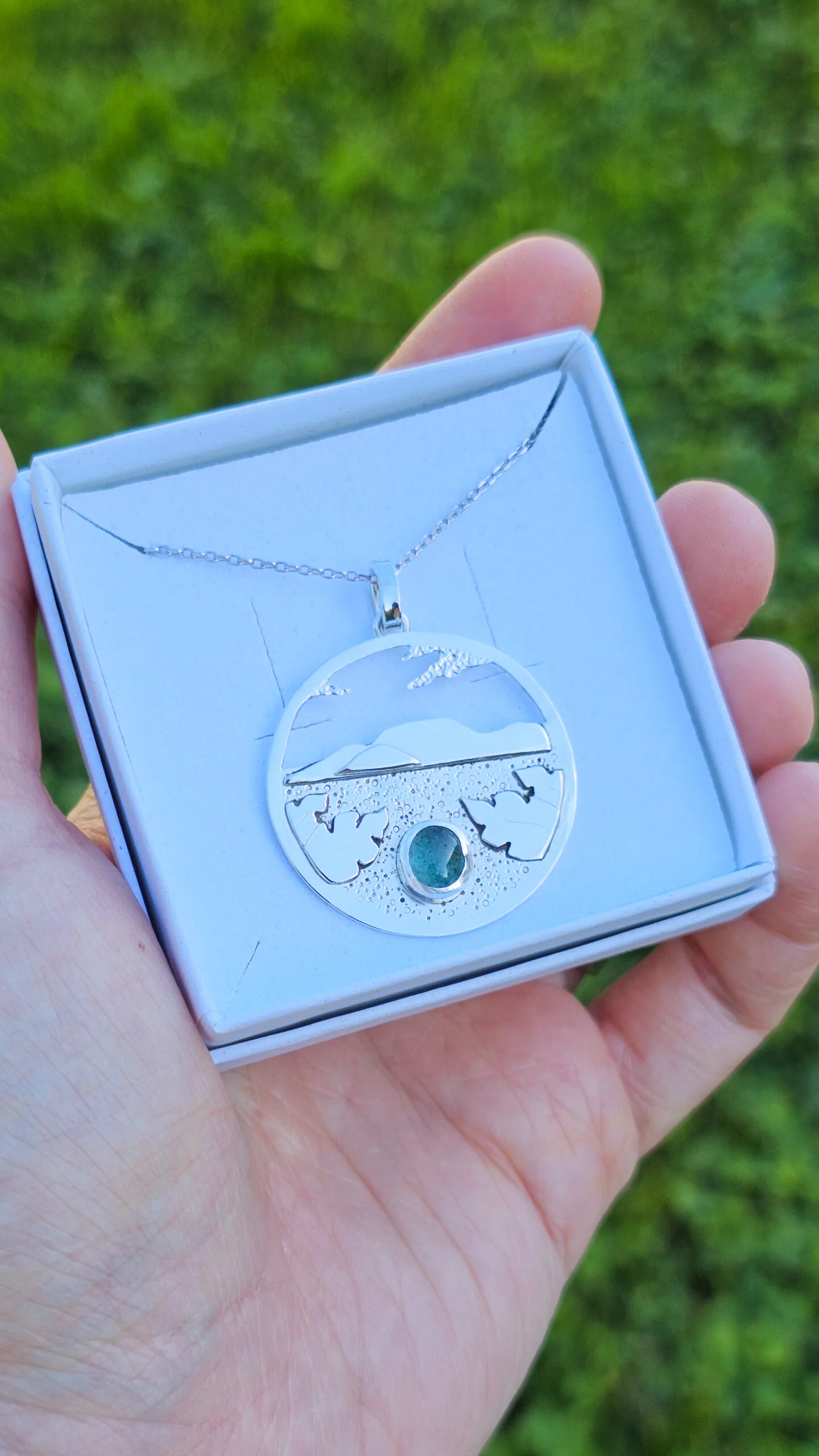 Collier en argent représentant un paysage tanzanien avec des montagnes et des feuilles de bananier, serti d'une tourmaline dichroïque bleue et verte