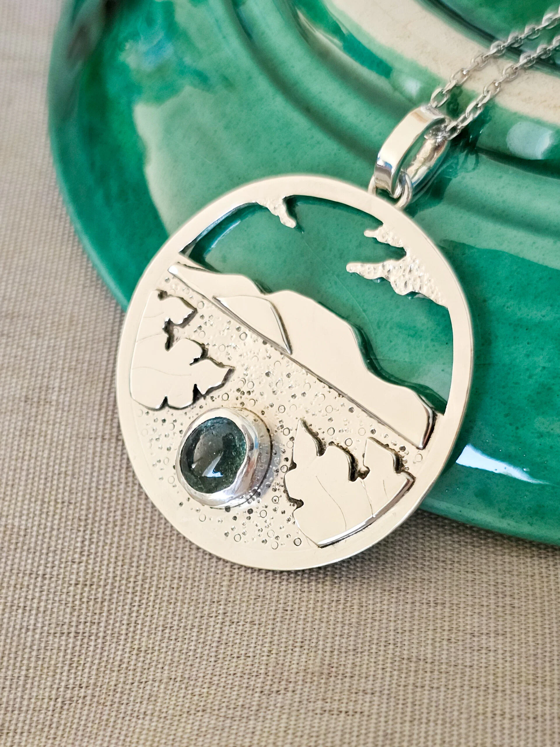 Collier en argent représentant un paysage tanzanien avec des montagnes et des feuilles de bananier, serti d'une tourmaline dichroïque bleue et verte